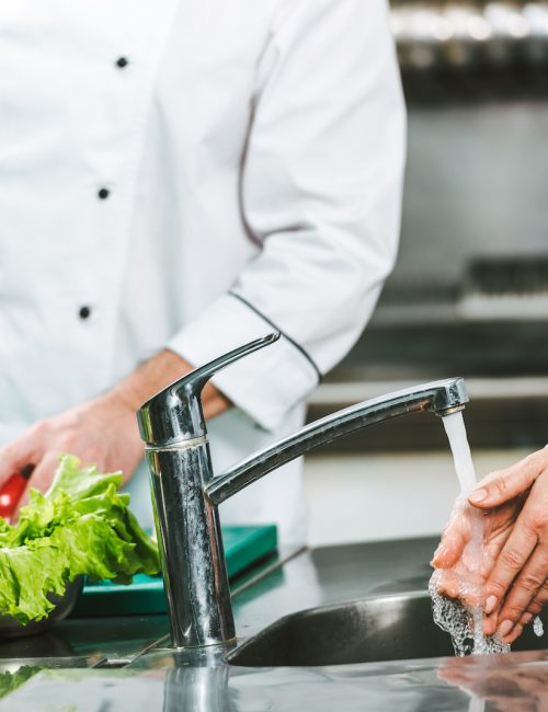 cropped view of female chef washing hands over sink while colleague cooking on background in restaurant kitchen